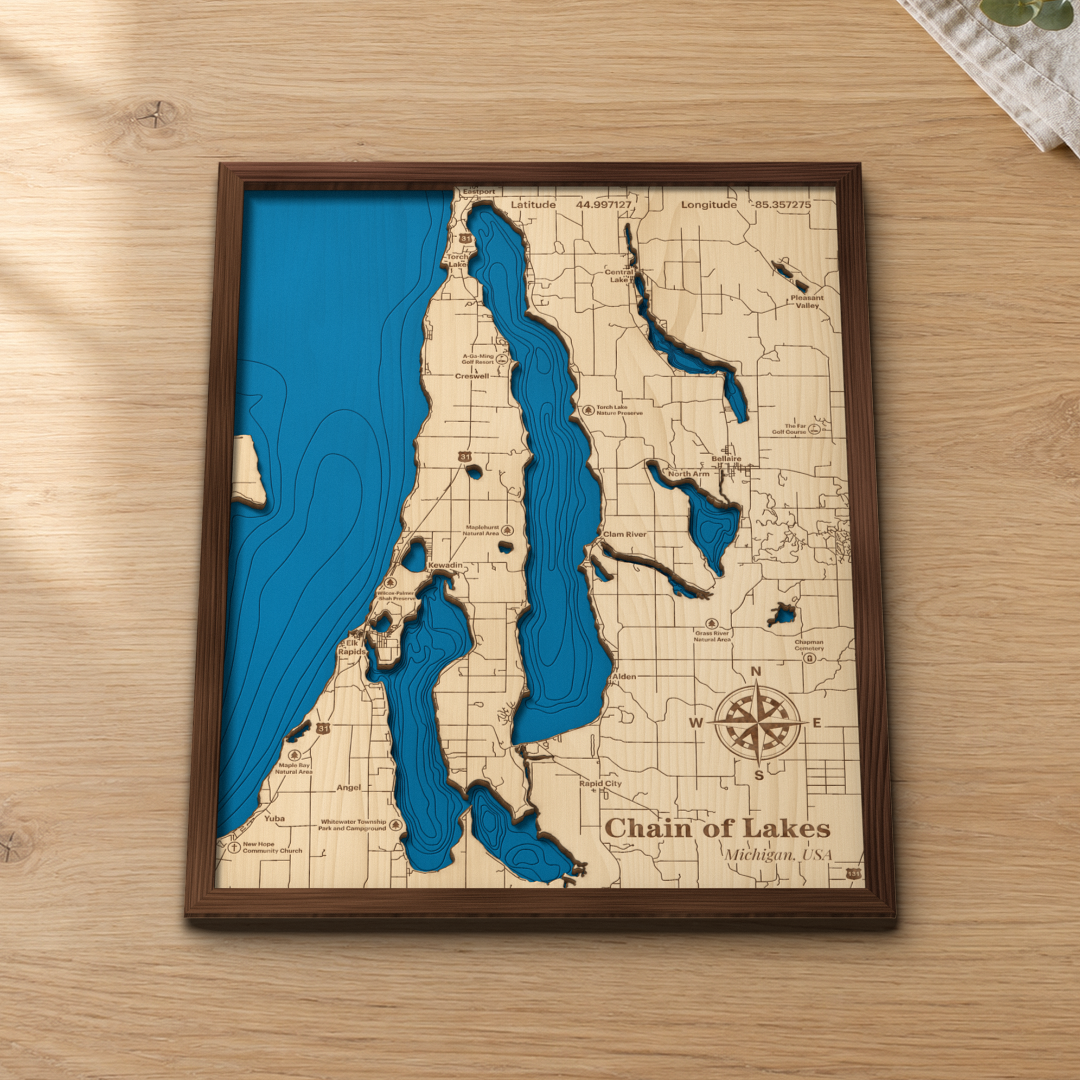 Chain of Lakes wood map lying flat on a wooden tabletop, showing the chain's depth contours from Torch Lake to Elk Rapids, with engraved towns, preserves and highways