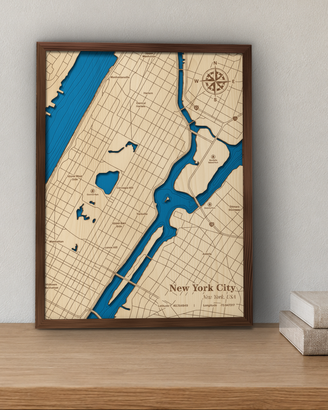 New York City wood map in a walnut frame standing on a wooden shelf against a wall beside a stack of books for scale